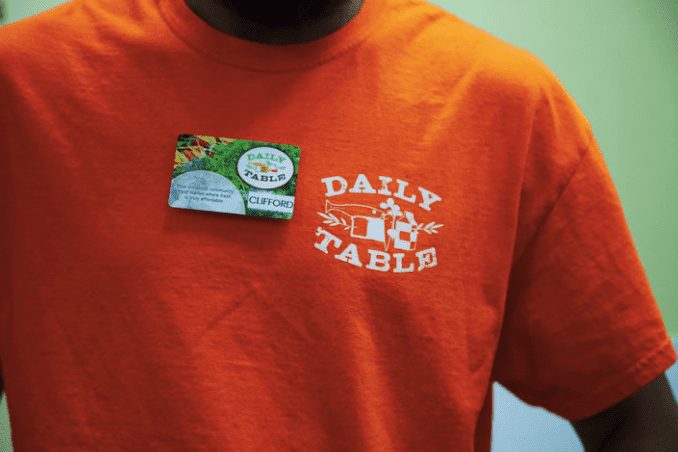 Close shot of an orange shirt with a white logo that reads Daily Table