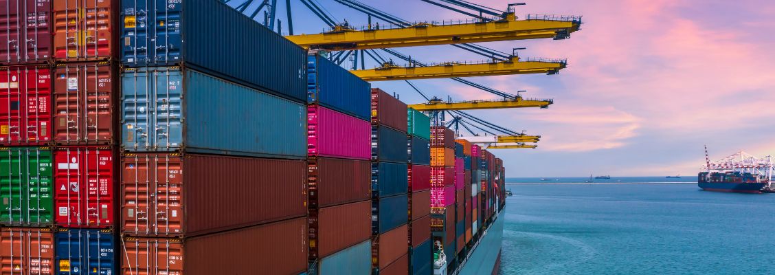 Cargo ship carrying shipping containers with cranes in the background. The sky is pink and blue close to sunset hour.