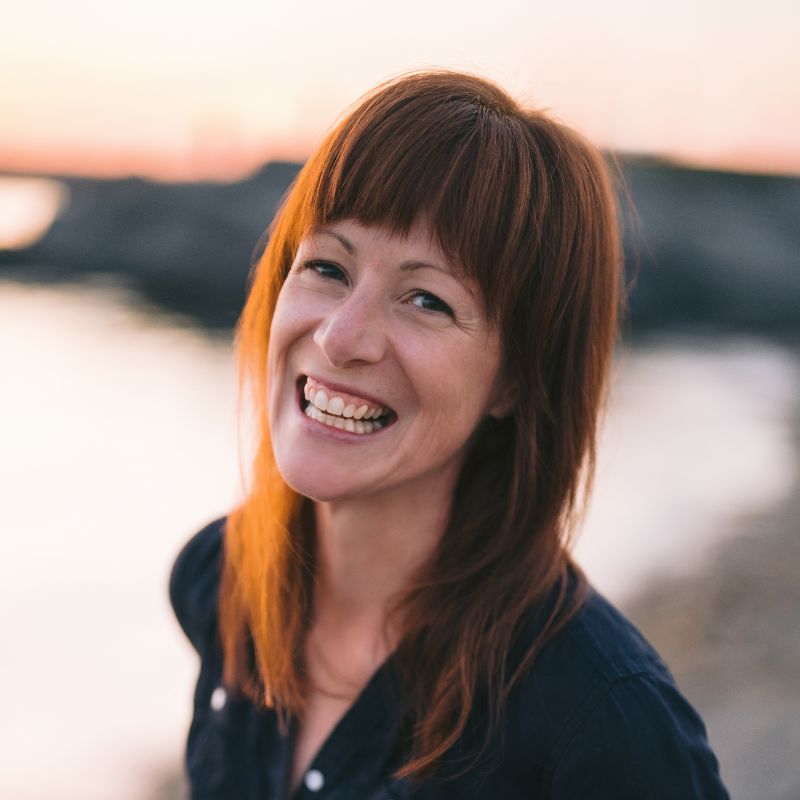 Headshot of white woman with long here and bangs smiling at camera. Blurry background