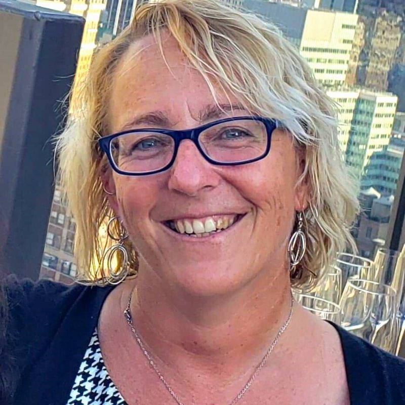 Headshot of blonde white woman looking and smiling at camera. She's wearing black frame glasses and a black cardigan