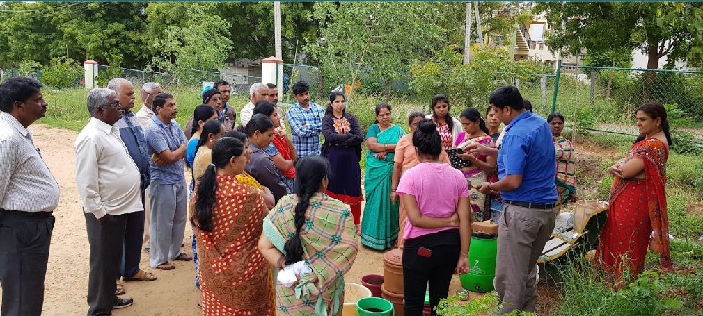 Group of people, Indian women and men in circle listening to man in blue shirt talking. They're outdoors in a green space.