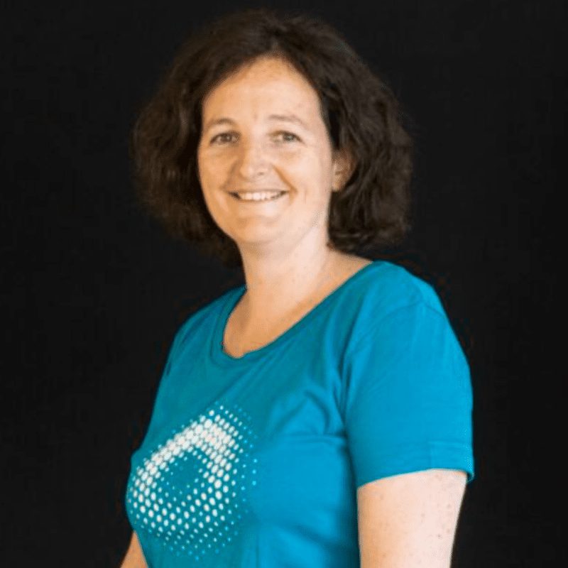 Portrait of a white woman smailing at camera in front of a black background. She's wearing a bright blue short sleeves t-shirt