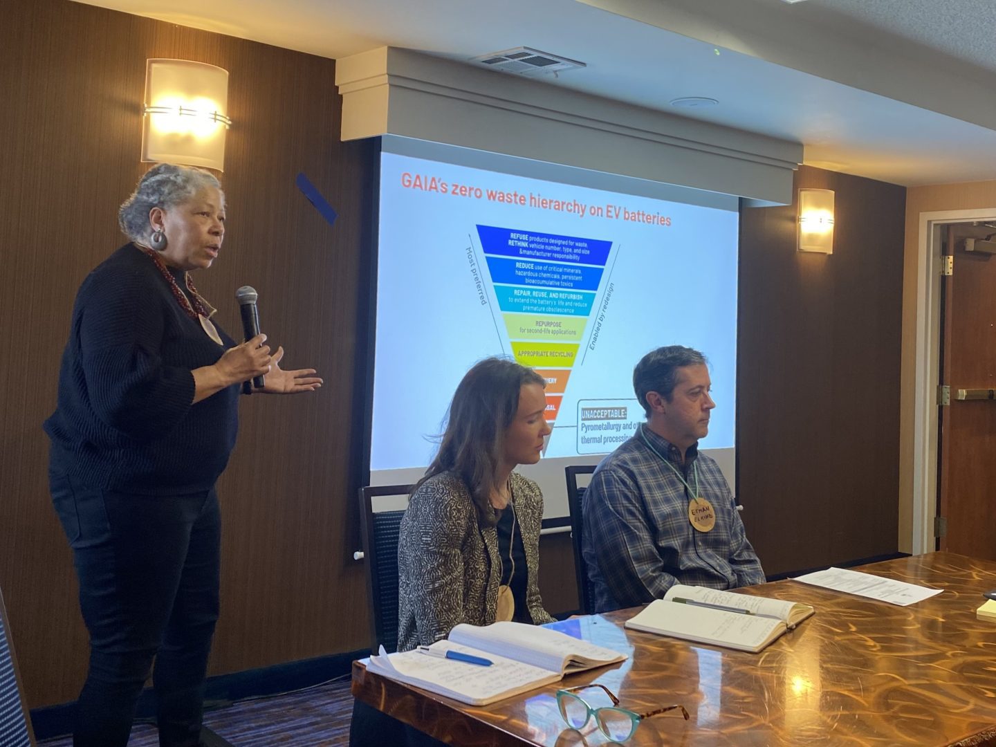 Panel of three people seating in front of a projection screen. Black woman dressed in black is holding a microphone