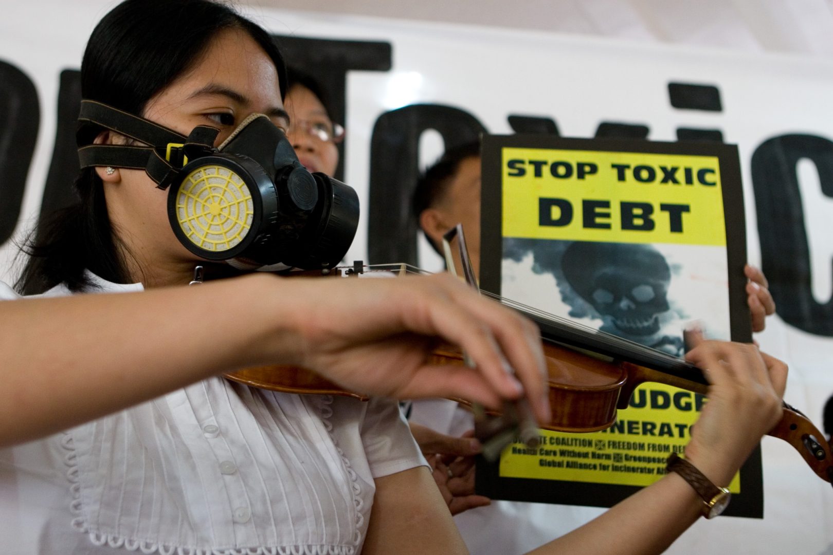 woman playing the violin wearing a gas mask, yellow sign in background says